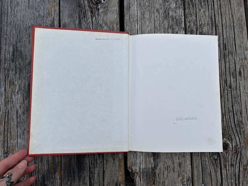 May include: An open hardcover book with a red spine and white endpapers. The left page features small black text reading 'Mullins School District No. 15 Library' at the top. The right page has the word 'DISCARDED' stamped in black ink.