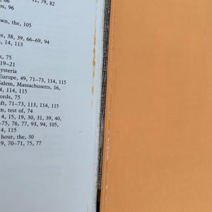 以下が含まれることがあります： オレンジ色の表紙と白いページを持つ本。ページにはセーラム、マサチューセッツ、ヒステリーなどの項目を含む索引が印刷されている。製本部分には露出した白いメッシュ生地と、黄色い接着剤の跡が縦に細く残っているのが見える。