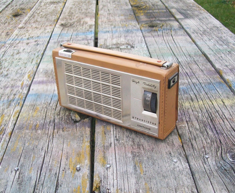 Może przedstawiać: Prostokątne przenośne radio tranzystorowe z obudową w kolorze opalenizny przypominającą sk&oacute;rę oraz srebrnym metalowym panelem przednim. Urządzenie posiada kratkę głośnika, pokrętło strojenia oraz nadruk High Fidelity, 8 Transistor i Oritone.