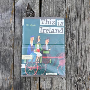 Peut inclure: Un livre reli&eacute; intitul&eacute; This is Ireland de M. Sasek avec une couverture vert fonc&eacute; illustrant trois personnes dans une cal&egrave;che. Le livre pr&eacute;sente une &eacute;tiquette d'&eacute;dition de biblioth&egrave;que Macmillan et du texte imprim&eacute; en blanc.