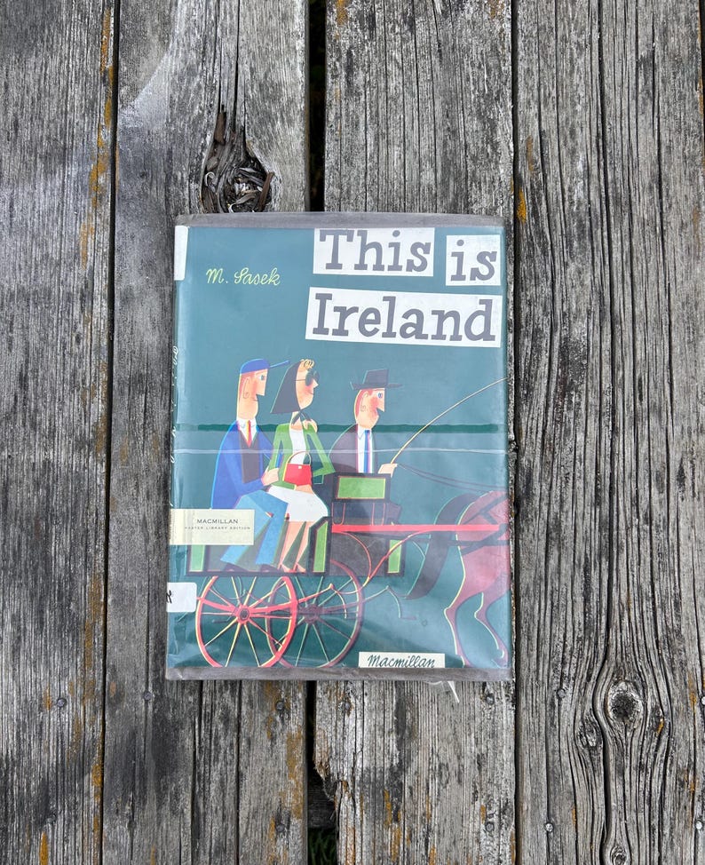 Peut inclure: Un livre reli&eacute; intitul&eacute; This is Ireland de M. Sasek avec une couverture vert fonc&eacute; illustrant trois personnes dans une cal&egrave;che. Le livre pr&eacute;sente une &eacute;tiquette d'&eacute;dition de biblioth&egrave;que Macmillan et du texte imprim&eacute; en blanc.