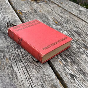 May include: A red hardcover book titled Roy Blakeley in the Haunted Camp by Percy Keese Fitzhugh. The front cover features the title in black text, and the spine displays the author's name and the publisher, Grosset & Dunlap, in black lettering.