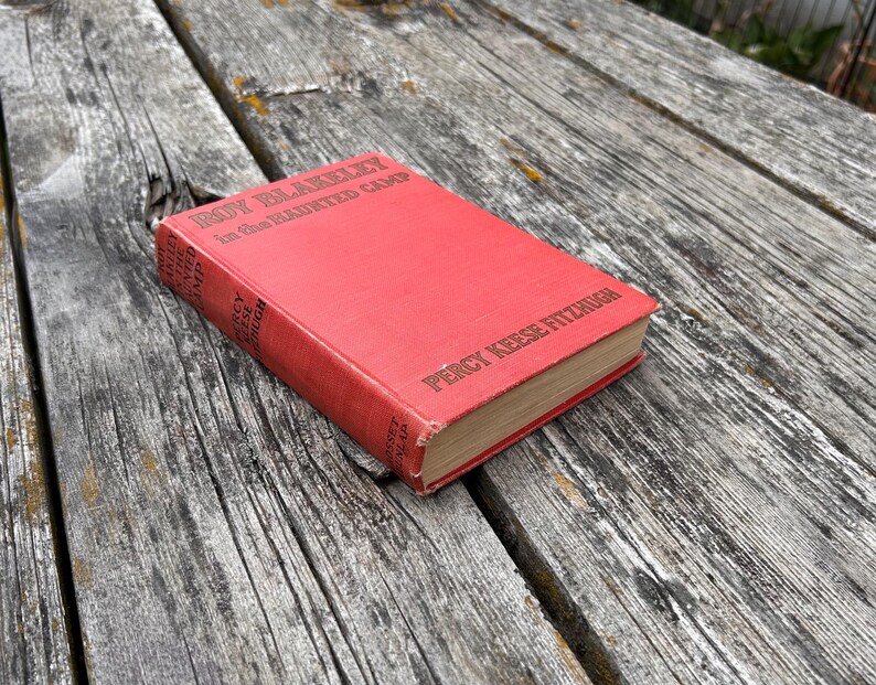 May include: A red hardcover book titled Roy Blakeley in the Haunted Camp by Percy Keese Fitzhugh. The front cover features the title in black text, and the spine displays the author's name and the publisher, Grosset & Dunlap, in black lettering.