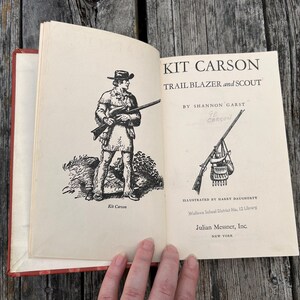 May include: An open hardcover book titled Kit Carson Trail Blazer and Scout by Shannon Garst. The left page features a black and white illustration of a scout, while the right page displays the title, author, and a rifle illustration.