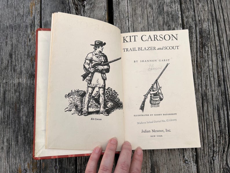 May include: An open hardcover book titled Kit Carson Trail Blazer and Scout by Shannon Garst. The left page features a black and white illustration of a scout, while the right page displays the title, author, and a rifle illustration.