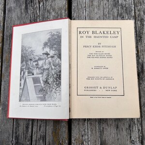 May include: An open hardcover book titled Roy Blakeley in the Haunted Camp by Percy Keese Fitzhugh. The left page features a black and white illustration of scouts, while the right page displays the title, author, and publisher Grosset & Dunlap.