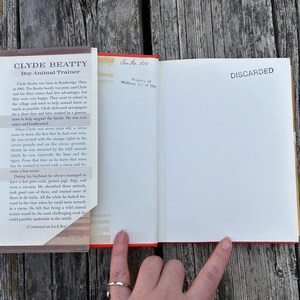 May include: An open hardcover book titled Clyde Beatty Boy Animal Trainer with a dust jacket flap on the left. The right page is blank with the word DISCARDED stamped in black ink. A small handwritten note reads Acc No 510 at the top.