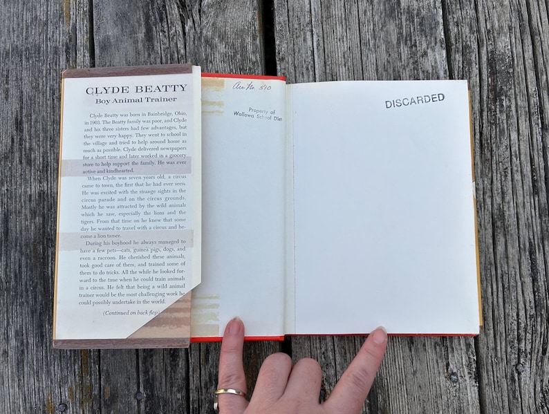 May include: An open hardcover book titled Clyde Beatty Boy Animal Trainer with a dust jacket flap on the left. The right page is blank with the word DISCARDED stamped in black ink. A small handwritten note reads Acc No 510 at the top.