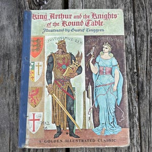Może przedstawiać: Książka w twardej oprawie zatytułowana King Arthur and the Knights of the Round Table, z ilustracjami Gustafa Tenggrena. Okładka przedstawia kr&oacute;la z mieczem i kobietę z sokołem, obok herb&oacute;w oraz napisu A Golden Illustrated Classic.