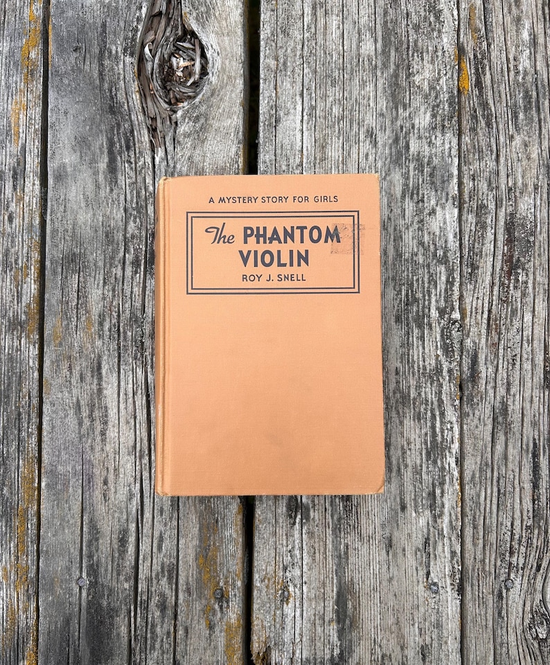 K&ouml;nnte beinhalten: Ein gebundenes Buch mit dem Titel The Phantom Violin von Roy J. Snell und dem Untertitel A Mystery Story for Girls. Das Buch hat einen einfarbigen, hellbraunen Einband mit schwarzem Text in einem rechteckigen Rahmen auf der Vorderseite.