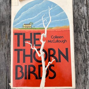 May include: A paperback book titled The Thorn Birds by Colleen McCullough. The cover features a white, leafless tree against a bright red background, with a blue arched sky and a yellow house in the distance. The title is printed in large black letters.