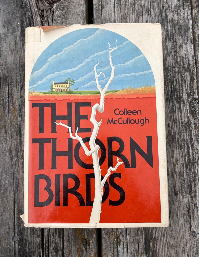 May include: A paperback book titled The Thorn Birds by Colleen McCullough. The cover features a white, leafless tree against a bright red background, with a blue arched sky and a yellow house in the distance. The title is printed in large black letters.