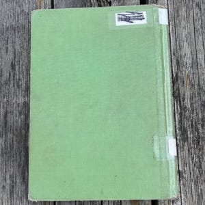 K&ouml;nnte beinhalten: Ein hellgr&uuml;nes Hardcover-Buch mit einem strukturierten Stoffeinband. Der Buchr&uuml;ckenbereich ist mit einem vertikal verlaufenden, transparenten Klebestreifen versehen. Oben befindet sich ein wei&szlig;er Bibliotheksaufkleber mit Barcode und Tinte.