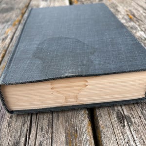 May include: A dark gray hardcover book with a textured fabric cover. The pages of the book are folded to create a distinct, symmetrical shape resembling a chalice or goblet visible along the fore-edge of the closed book's paper block.