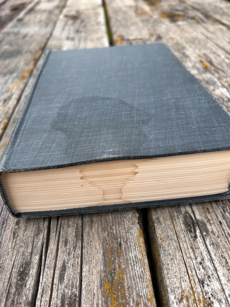 May include: A dark gray hardcover book with a textured fabric cover. The pages of the book are folded to create a distinct, symmetrical shape resembling a chalice or goblet visible along the fore-edge of the closed book's paper block.