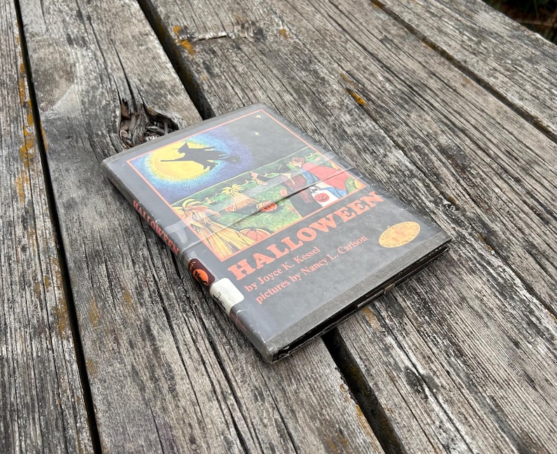 Może przedstawiać: Książka w twardej oprawie zatytułowana Halloween autorstwa Joyce K. Kessel z ilustracjami Nancy L. Carlson. Okładka przedstawia ciemną ilustrację czarownicy lecącej na tle dużego ż&oacute;łtego księżyca nad sceną z dyniami i postaciami w kostiumach.