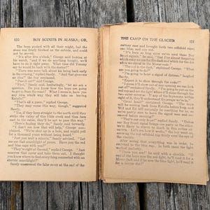 K&ouml;nnte beinhalten: Ein aufgeschlagenes altes Buch mit gealterten, vergilbten Seiten und schwarzem Drucktext. Die linke Seite tr&auml;gt den Titel Boy Scouts in Alaska; Or, und die rechte Seite The Camp on the Glacier. Die Seiten weisen deutliche Gebrauchsspuren auf.