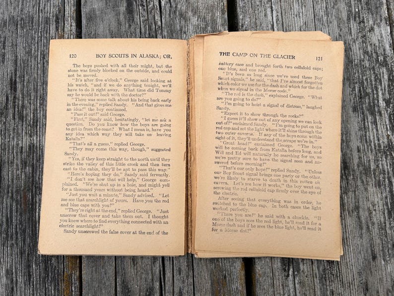 K&ouml;nnte beinhalten: Ein aufgeschlagenes altes Buch mit gealterten, vergilbten Seiten und schwarzem Drucktext. Die linke Seite tr&auml;gt den Titel Boy Scouts in Alaska; Or, und die rechte Seite The Camp on the Glacier. Die Seiten weisen deutliche Gebrauchsspuren auf.