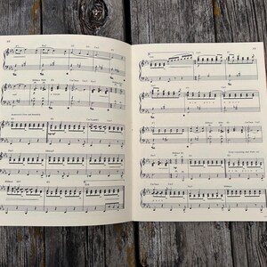 K&ouml;nnte beinhalten: Ein aufgeschlagenes Buch mit gedruckten Noten, das schwarze Notensysteme, Noten und Akkordsymbole auf cremefarbenem Papier zeigt. Die Partitur enth&auml;lt mehrere Zeilen Klaviermusik mit verschiedenen Rhythmen, Dynamik und Tempoangaben.