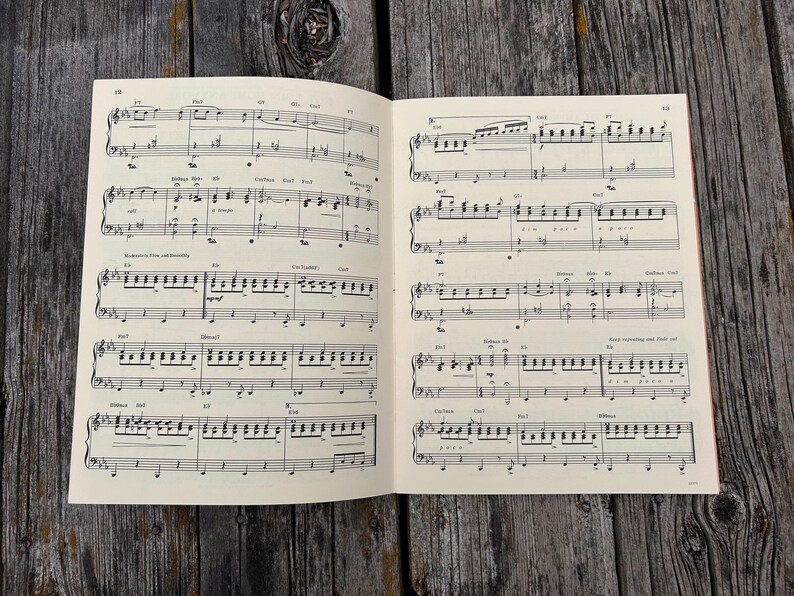K&ouml;nnte beinhalten: Ein aufgeschlagenes Buch mit gedruckten Noten, das schwarze Notensysteme, Noten und Akkordsymbole auf cremefarbenem Papier zeigt. Die Partitur enth&auml;lt mehrere Zeilen Klaviermusik mit verschiedenen Rhythmen, Dynamik und Tempoangaben.