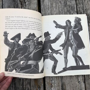 Puede incluir: Un libro de tapa dura abierto con ilustraciones hist&oacute;ricas en blanco y negro de soldados con uniformes militares del siglo XVIII. El texto describe la Revoluci&oacute;n Americana, incluyendo las frases, A tub for the whale, y Mutinous soldiers force Congress to flee from Philadelphia.
