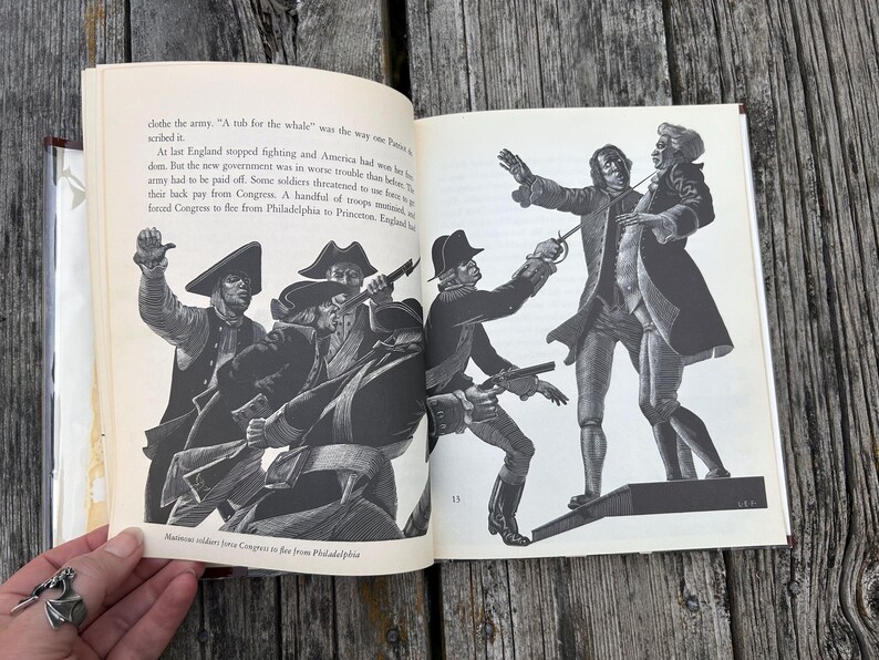 Puede incluir: Un libro de tapa dura abierto con ilustraciones hist&oacute;ricas en blanco y negro de soldados con uniformes militares del siglo XVIII. El texto describe la Revoluci&oacute;n Americana, incluyendo las frases, A tub for the whale, y Mutinous soldiers force Congress to flee from Philadelphia.