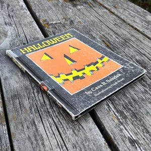 May include: A hardback book titled Halloween by Cass R. Sandak with illustrations by Frank Bozzi. The black cover features a large orange jack-o'-lantern graphic with yellow triangular eyes and a jagged yellow mouth centred on the front panel.