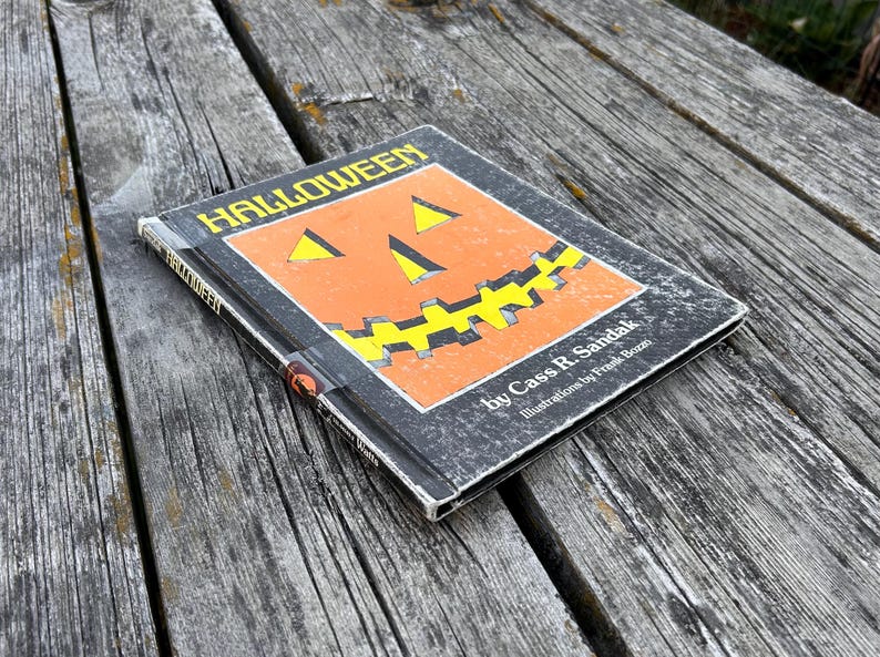 May include: A hardback book titled Halloween by Cass R. Sandak with illustrations by Frank Bozzi. The black cover features a large orange jack-o'-lantern graphic with yellow triangular eyes and a jagged yellow mouth centred on the front panel.