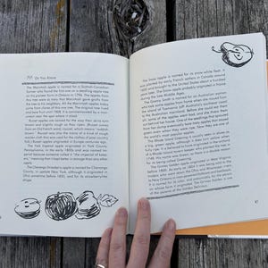 以下が含まれることがあります： 白いページに黒い文字が印刷され、手描きのリンゴのイラストが添えられた開いた本。左ページには86、右ページには87の番号が振られている。テキストには、さまざまなリンゴの品種に関する歴史的な事実が記載されている。