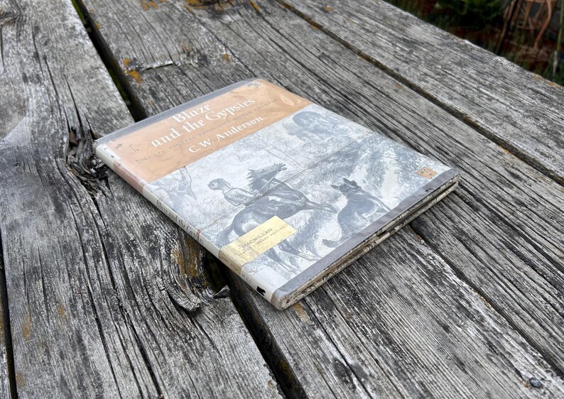 May include: A hardcover book titled Blaze and the Gypsies by C.W. Anderson. The cover features a black and white illustration of a person riding a horse with a dog nearby. The top section is tan, and a small yellow library sticker is on the bottom.