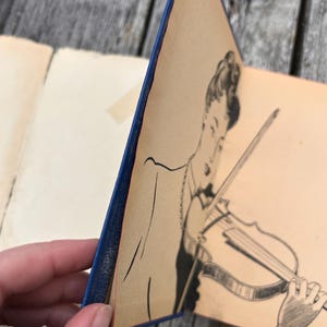 May include: A vintage book with a blue spine and a cream-coloured page featuring a black ink illustration of a person playing a violin. The drawing depicts the individual from the chest up, holding the instrument with a bow, rendered in fine lines.