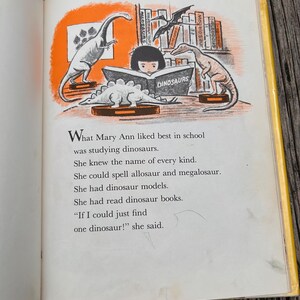 Pode incluir: Uma p&aacute;gina de um livro infantil com uma ilustra&ccedil;&atilde;o de uma crian&ccedil;a lendo um livro intitulado DINOSAURS, cercada por modelos de dinossauros em prateleiras e mesas. O texto diz: What Mary Ann liked best in school was studying dinosaurs.