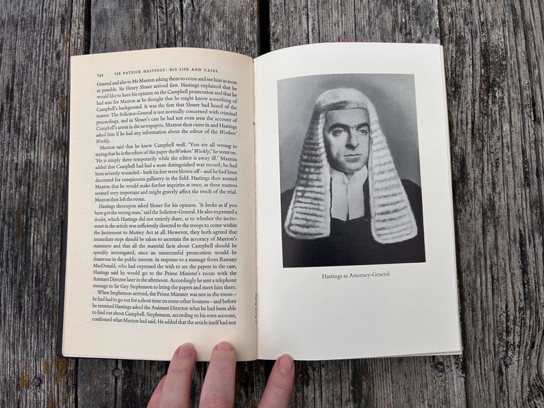 Pu&ograve; includere: Un libro aperto con pagine color crema, testo nero e un ritratto in bianco e nero di una persona che indossa una parrucca e una toga legale tradizionale. La didascalia sotto il ritratto recita: "Hastings as Attorney-General".