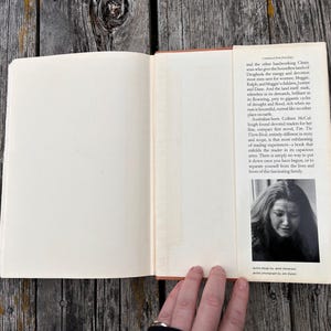 May include: An open hardcover book with a cream-colored dust jacket flap. The flap contains printed text describing the novel The Thorn Birds and includes a black and white portrait photograph of the author, Colleen McCullough, at the bottom.