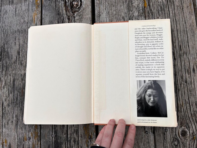 May include: An open hardcover book with a cream-colored dust jacket flap. The flap contains printed text describing the novel The Thorn Birds and includes a black and white portrait photograph of the author, Colleen McCullough, at the bottom.