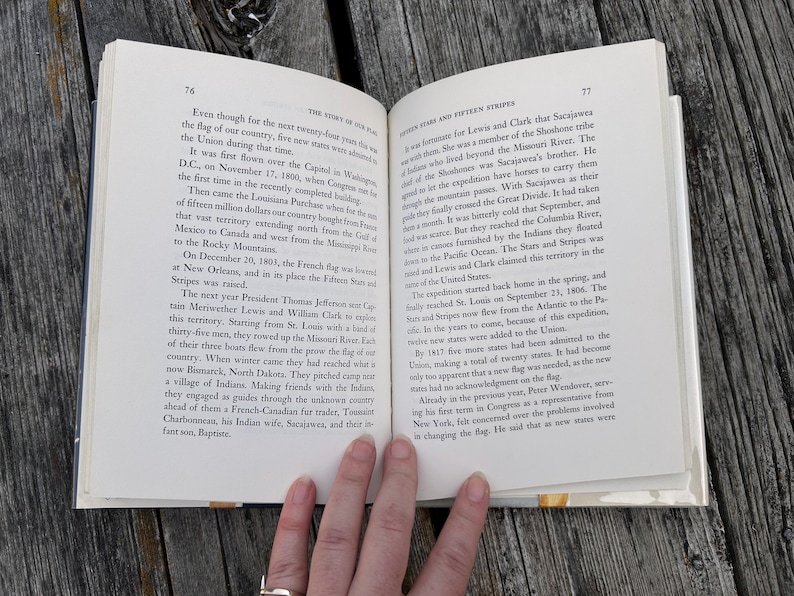 May include: An open hardcover book with white pages and black printed text. The left page is numbered 76 with the header The Story of Our Flag. The right page is numbered 77 with the header Fifteen Stars and Fifteen Stripes.