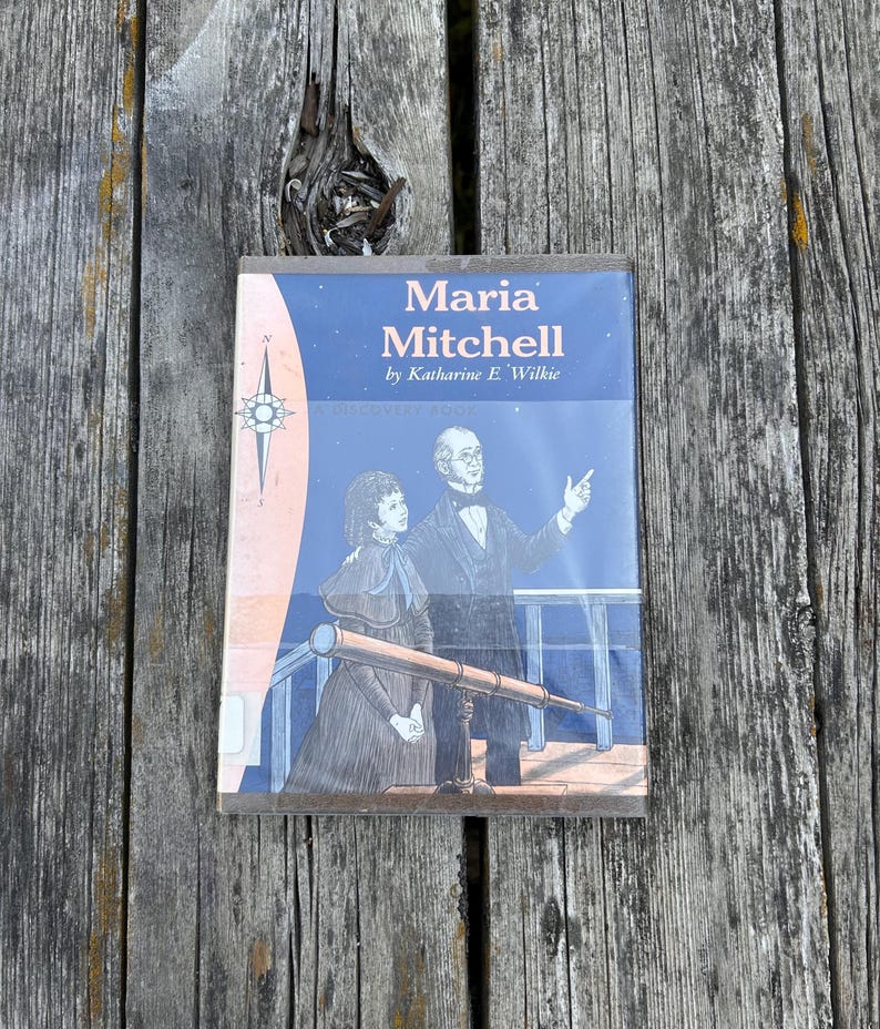 Puede incluir: Un libro biogr&aacute;fico titulado Maria Mitchell de Katharine E. Wilkie. La portada presenta una ilustraci&oacute;n azul de una mujer y un hombre en una terraza con un telescopio, una rosa de los vientos y un cielo nocturno estrellado.