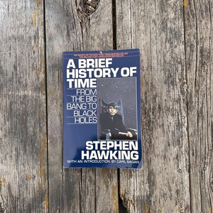 Peut inclure: Livre &laquo;&nbsp;Une br&egrave;ve histoire du temps&nbsp;&raquo; de Stephen Hawking. La couverture est bleu fonc&eacute; avec du texte blanc et une photo de Stephen Hawking. Le livre est sur une surface en bois vieilli.