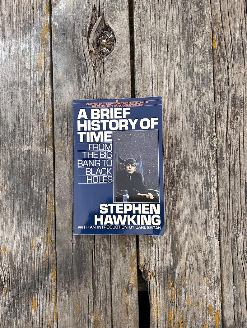 Peut inclure: Livre &laquo;&nbsp;Une br&egrave;ve histoire du temps&nbsp;&raquo; de Stephen Hawking. La couverture est bleu fonc&eacute; avec du texte blanc et une photo de Stephen Hawking. Le livre est sur une surface en bois vieilli.