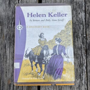 May include: A hardback book titled Helen Keller by Stewart and Polly Anne Graff, A Discovery Book. The cover features a purple and yellow illustration of a person riding a horse and another person standing nearby, illustrated by Paul Frame.