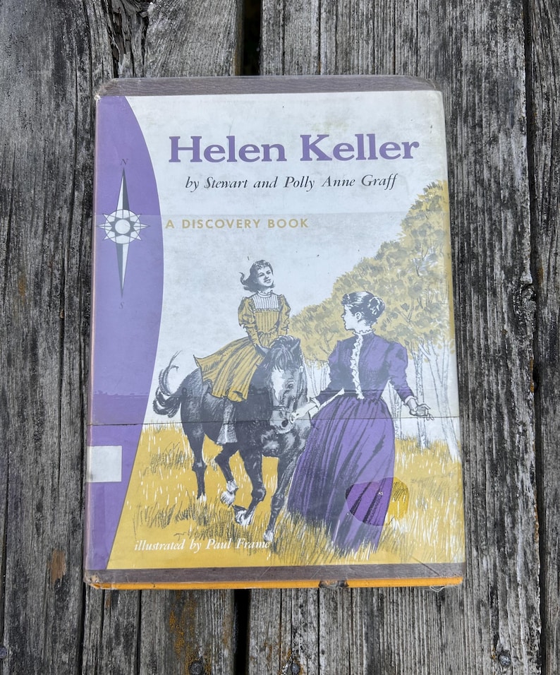 May include: A hardback book titled Helen Keller by Stewart and Polly Anne Graff, A Discovery Book. The cover features a purple and yellow illustration of a person riding a horse and another person standing nearby, illustrated by Paul Frame.
