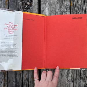 May include: A hardcover book titled The Gobble-uns'll Git You Ef You Don't Watch Out! by James Whitcomb Riley, illustrated by Joel Schick. The book features a white dust jacket flap with red text and a bright red endpaper with the word DISCARDED printed.