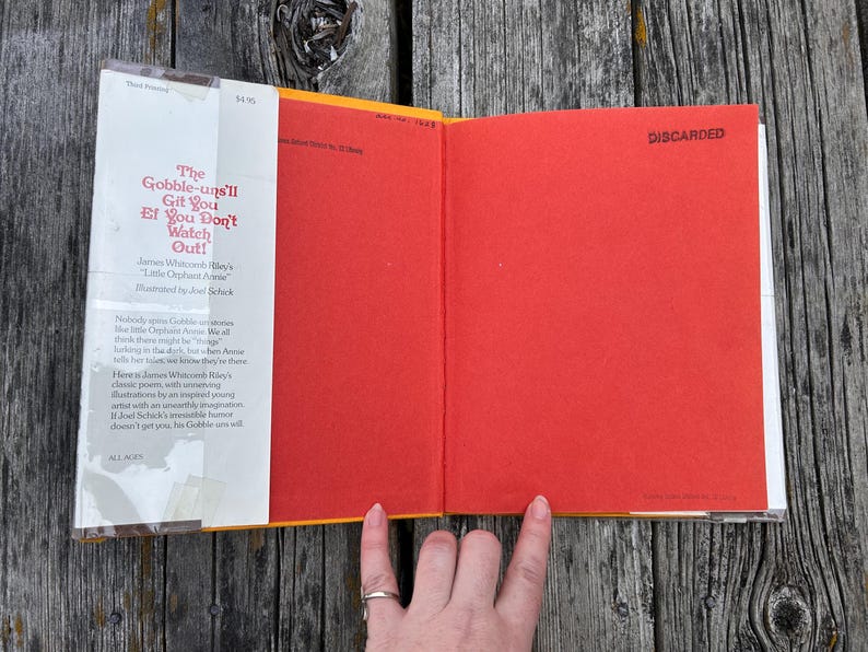 May include: A hardcover book titled The Gobble-uns'll Git You Ef You Don't Watch Out! by James Whitcomb Riley, illustrated by Joel Schick. The book features a white dust jacket flap with red text and a bright red endpaper with the word DISCARDED printed.