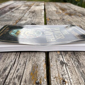 K&ouml;nnte beinhalten: Ein Taschenbuch mit dem Titel The Power of Myth von Joseph Campbell mit einem schwarzen Einband und einem kreisf&ouml;rmigen Grafikdesign. Das Buch ist aus einer niedrigen Perspektive abgebildet, die die wei&szlig;en Seiten und die gl&auml;nzende Oberfl&auml;che des Covers zeigt.