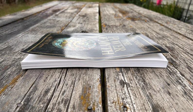 K&ouml;nnte beinhalten: Ein Taschenbuch mit dem Titel The Power of Myth von Joseph Campbell mit einem schwarzen Einband und einem kreisf&ouml;rmigen Grafikdesign. Das Buch ist aus einer niedrigen Perspektive abgebildet, die die wei&szlig;en Seiten und die gl&auml;nzende Oberfl&auml;che des Covers zeigt.