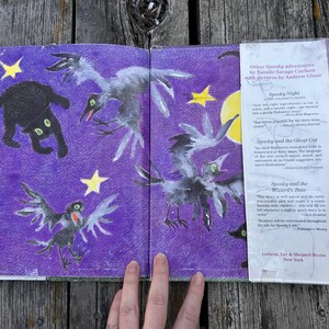 K&ouml;nnte beinhalten: Ein aufgeschlagenes Kinderbuch mit violettem Hintergrund, das eine schwarze Katze, drei graue V&ouml;gel und gelbe Sterne zeigt. Die rechte Seite enth&auml;lt eine wei&szlig;e Schutzumschlagklappe mit Text, der weitere Spooky-Abenteuer von Natalie Savage Carlson auflistet.