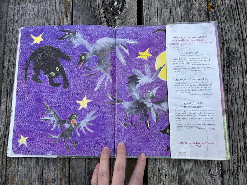K&ouml;nnte beinhalten: Ein aufgeschlagenes Kinderbuch mit violettem Hintergrund, das eine schwarze Katze, drei graue V&ouml;gel und gelbe Sterne zeigt. Die rechte Seite enth&auml;lt eine wei&szlig;e Schutzumschlagklappe mit Text, der weitere Spooky-Abenteuer von Natalie Savage Carlson auflistet.