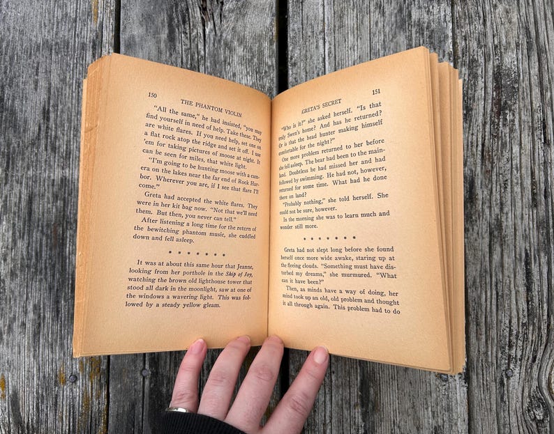 K&ouml;nnte beinhalten: Ein aufgeschlagenes Buch mit gealterten, hellbraunen Seiten und schwarzem Drucktext. Die linke Seite tr&auml;gt den Titel The Phantom Violin und die rechte Seite den Titel Greta's Secret. Das Buch ist ge&ouml;ffnet und zeigt zwei volle Textseiten.