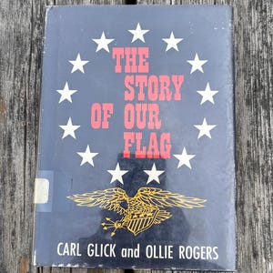 May include: A hardcover book titled The Story of Our Flag by Carl Glick and Ollie Rogers. The dark blue cover features a circle of white stars surrounding the red text, with a gold eagle illustration holding arrows at the bottom.