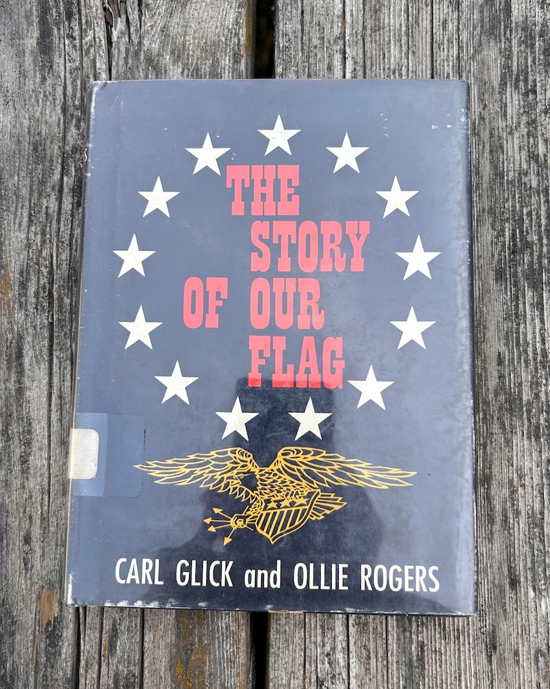 May include: A hardcover book titled The Story of Our Flag by Carl Glick and Ollie Rogers. The dark blue cover features a circle of white stars surrounding the red text, with a gold eagle illustration holding arrows at the bottom.